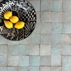 Op de afbeelding: Een decoratieve zwart-wit patroonkom gevuld met vijf gele citroenen. De kom staat op een vierkant, lichtblauw betegeld oppervlak. Het ontwerp van de kom omvat geometrische en bloemmotieven.