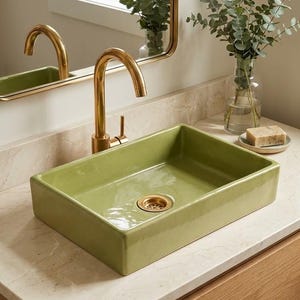 May include: A rectangular sage green ceramic vessel sink with a glossy finish and a gold-toned metal faucet and drain. The sink is mounted on a light-colored stone countertop, accompanied by a clear glass vase with green eucalyptus stems.