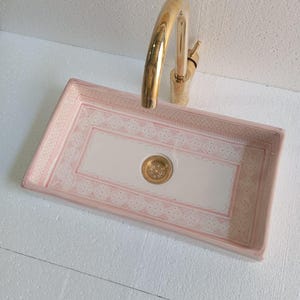 May include: A rectangular ceramic sink with a pink and white decorative pattern and a gold faucet. The sink has a pink border with a white center and a gold drain. The faucet is curved and has a gold finish.