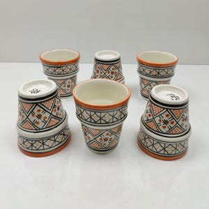 Könnte beinhalten: Sechs Keramikbecher mit traditionellem marokkanischem Design. Die Becher haben einen weißen Sockel mit orangefarbenen, schwarzen und weißen geometrischen Mustern. Jeder Becher hat einen orangefarbenen Rand und eine einzigartige Form.