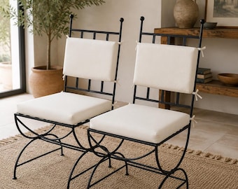 Op de afbeelding: Twee stoelen met zwarte metalen frames, gebogen poten en een rugleuning met een rasterpatroon. Elke stoel is voorzien van een afneembaar gebroken wit zitkussen en een bijpassend rechthoekig rugkussen, beide vastgezet met dunne stoffen linten.