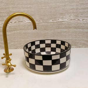 May include: A gold-colored faucet with cross handles stands next to a black and white checkered ceramic bowl. The bowl is round with a wide rim and a glossy finish. The faucet has a curved spout.
