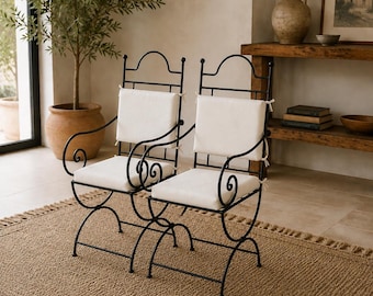 Op de afbeelding: Twee zwarte metalen stoelen met gebogen armleuningen, gewelfde rugleuningen en vierkante gebroken witte zitkussens. De stoelen hebben een frame van dunne metalen staven met decoratieve kruldetails bij de armleuningen en de basis.