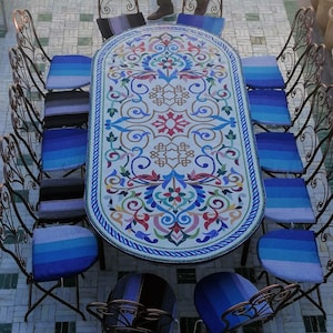 May include: A white oval table with a colourful mosaic design and a blue border. The table is surrounded by 10 black metal chairs with blue and white striped cushions.