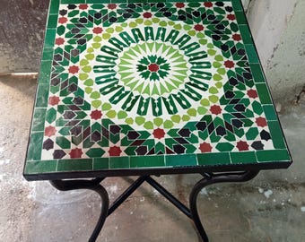 Puede incluir: Mesa cuadrada de mosaico con base de metal negro. La encimera presenta un patrón geométrico en tonos verdes, rojos y blancos. La mesa mide aproximadamente 60 cm cuadrados y es adecuada para uso en interiores o exteriores.