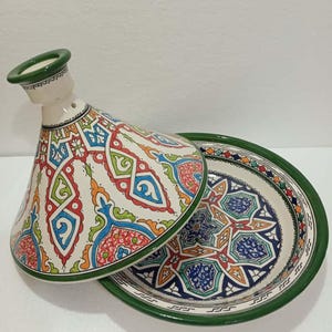 May include: A ceramic tagine with a conical lid and a matching base. The lid and base are white with intricate, colorful geometric patterns in red, blue, green, and orange. The rim of the base and the top of the lid are green.