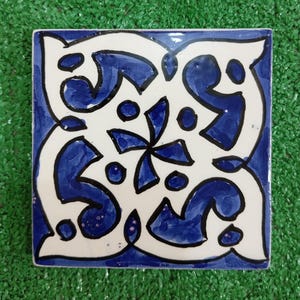 May include: A square ceramic tile with a blue and white geometric pattern. The pattern features a central flower with four petals and a swirling design around the edges.