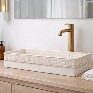 Puede incluir: Un lavabo rectangular de cerámica blanquecina con un borde decorativo. Un grifo dorado está montado en la pared. Una toalla blanca doblada descansa sobre la encimera de madera. También se ven una planta en maceta y un dispensador de jabón.