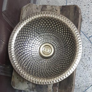 Può includere: Un lavandino rotondo in ottone martellato con uno scarico circolare al centro. Il lavandino ha una finitura martellata testurizzata ed è di colore oro chiaro.