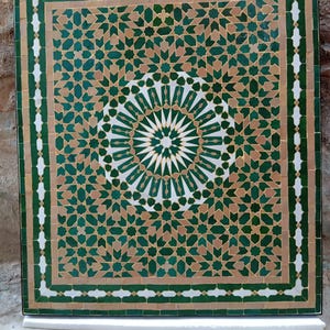 May include: Square mosaic tile with a geometric pattern in shades of green, tan, and white. The design features a central starburst pattern surrounded by intricate shapes and borders. The tile is framed with a black border.