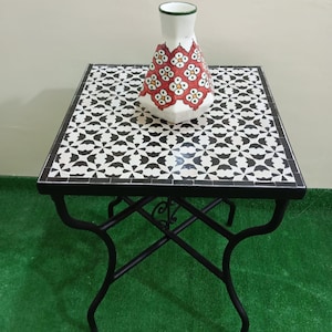 May include: A black metal side table with a square top made of white and black mosaic tiles. A white ceramic vase with red and green floral designs sits on the table.