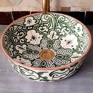 以下が含まれることがあります： 緑と白の花柄が特徴の丸いセラミック洗面台。洗面台は銅色の縁と金色の排水口が付いています。複雑な花柄が洗面台の表面全体を覆っています。