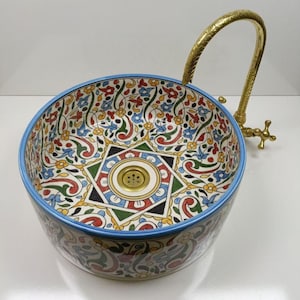 May include: A patterned ceramic sink with a blue rim and a gold-coloured tap. The sink features a colourful floral design in red, blue, yellow, and green. The sink's design includes a central star-shaped motif.