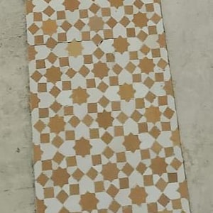 Puede incluir: Una baldosa rectangular con un patrón repetitivo de estrellas de ocho puntas y pequeños cuadrados en tonos beige y blanco. El diseño es simétrico y cubre toda la superficie.