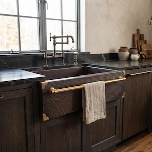 Könnte beinhalten: Ein dunkelbraunes, gehämmertes Kupfer-Spülbecken im Landhausstil mit Messing-Eckakzenten und einer horizontalen Messing-Handtuchstange. Ein beiges Leinentuch hängt an der Stange. Das Spülbecken ist in eine schwarze Marmorarbeitsplatte eingelassen.