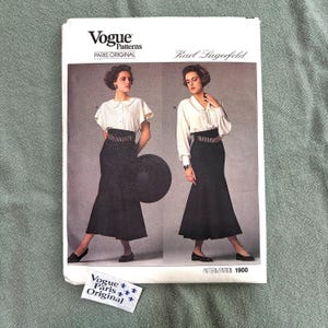 May include: A Vogue Patterns Paris Original sewing pattern envelope for Karl Lagerfeld design 1900. The white envelope features two illustrations of a person wearing a white blouse and a black high-waisted skirt with a wide, textured belt.