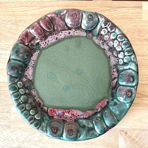 May include: A round ceramic plate featuring a smooth, matte sage green center with small teal speckles. The wide, textured rim is decorated with raised, rounded organic shapes in shades of deep burgundy, forest green, and metallic teal glaze.