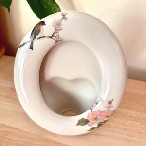Puede incluir: Un marco de fotos ovalado de cerámica blanca con un diseño pintado a mano de un pequeño pájaro marrón y blanco posado en una rama floreciente con delicadas flores rosas y hojas verdes a lo largo del borde curvo, liso y brillante.