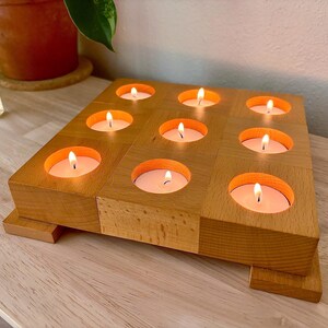 May include: A square light brown wooden candle holder featuring nine circular cutouts arranged in a grid pattern. Each cutout contains a small white tea light candle with a lit flame. The wooden base is elevated by two small rectangular feet.