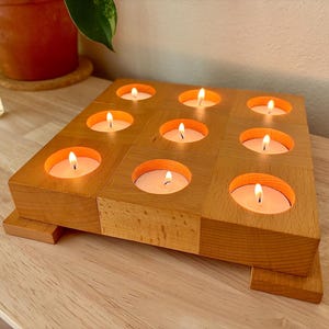 May include: A square light-colored wooden candle holder featuring nine circular cutouts arranged in a grid. Each cutout holds a small, lit white tea light candle. The wooden base is elevated by small rectangular feet at each corner.