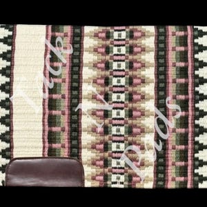 Może przedstawiać: Prostokątna podkładka pod siodło z tkanym wzorem w kolorze kremowym, różowym, zielonym i brązowym. Podkładka ma powtarzający się geometryczny wzór i kawałek ciemnobrązowej skóry na dole. Krawędzie wykończone są białą obwódką.