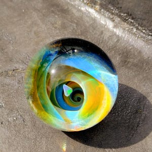 May include: A clear glass marble with an internal design of swirling blue, yellow, and green colors. The marble has a spiral pattern and a small, triangular, iridescent shape at its center. The marble is resting on a gray surface.