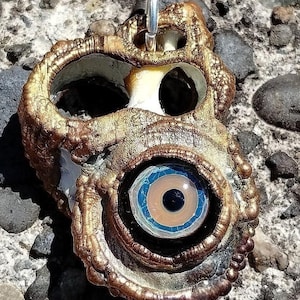 May include: A gold-toned pendant with a large, blue and white eye design. The pendant has a textured, organic shape and a silver loop for hanging.
