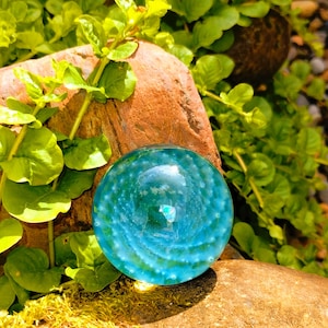 Könnte beinhalten: Eine klare, blaue Glaskugel mit einem Wirbelmuster und kleinen weißen Einschlüssen. Die Kugel liegt auf einem braunen Felsen, umgeben von grünem Laub und Moos. Das Design der Kugel ähnelt einer Unterwasserszene.