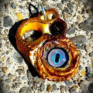 May include: A gold-colored seashell pendant with a colorful glass eye. The eye has a black center and concentric rings of blue, green, red, and yellow.