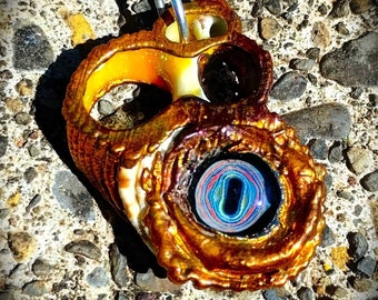 Puede incluir: Un colgante de concha dorada con un ojo de cristal de colores. El ojo tiene un centro negro y anillos concéntricos de azul, verde, rojo y amarillo.