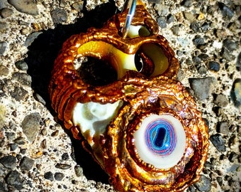 Puede incluir: Un colgante de color dorado con un diseño blanco en forma de ojo. El ojo tiene un iris azul y rosa y una pupila blanca. El colgante está hecho de metal y tiene una superficie texturizada.