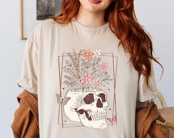 May include: A beige t-shirt with a graphic of a skull with flowers growing out of the top. The skull is outlined in brown and the flowers are pink, orange, and white.