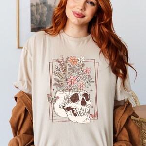May include: A beige t-shirt with a graphic of a skull with flowers growing out of the top. The skull is outlined in brown and the flowers are pink, orange, and white.