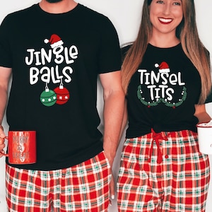 May include: Matching black Christmas t-shirts with humorous text and Santa hats. One shirt says "Jingle Balls" with ornament graphics, the other says "Tinsel Tits". Both people wear red plaid pajama pants and hold mugs.