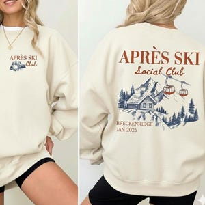 May include: Cream-colored sweatshirt with "APRÈS SKI Club" printed on the front and "APRÈS SKI Social Club" on the back. The back graphic features a cabin, mountains, and ski lifts. The text "BRECKENRIDGE JAN 2026" is also printed on the back.