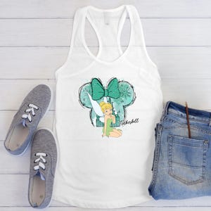 May include: White vest top featuring a graphic of Tinkerbell with a teal bow and Mickey Mouse ears. The top is styled with blue jeans and grey trainers.