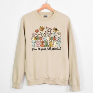 May include: A beige sweatshirt with the words "OCCUPATIONAL THERAPY" in colorful, retro-style lettering, above the phrase "grow to your full potential." The design includes a floral arrangement with yellow, orange, and white flowers.