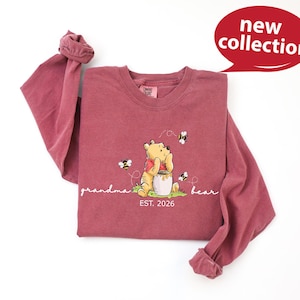 A red long-sleeved t-shirt with a Winnie the Pooh graphic and the text "Grandma Bear" printed on it, along with a speech bubble indicating a new collection.