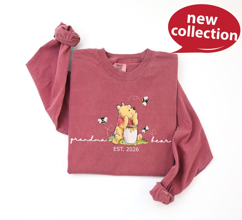 A red long-sleeved t-shirt with a Winnie the Pooh graphic and the text "Grandma Bear" printed on it, along with a speech bubble indicating a new collection.