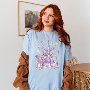 Puede incluir: Camiseta azul claro con una ilustraci&oacute;n colorida de un castillo y globos. El dise&ntilde;o del castillo utiliza tonos morados, rosas y amarillos. La camiseta est&aacute; hecha de un material suave.