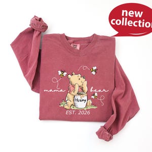 A pink long-sleeved t-shirt with a Winnie the Pooh character and honey pot design, along with the text "mama bear" and "Est. 2026".