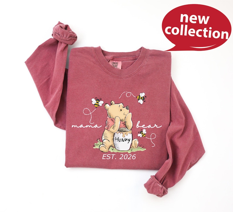 A pink long-sleeved t-shirt with a Winnie the Pooh character and honey pot design, along with the text "mama bear" and "Est. 2026".