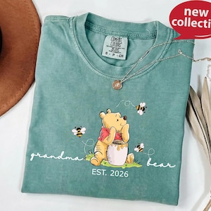 A green t-shirt with a Winnie the Pooh-themed design, featuring the character sitting on a grassy field with a honey pot and a bee nearby. The t-shirt also has the text "grandma bear" and "EST. 2026" printed on it.
