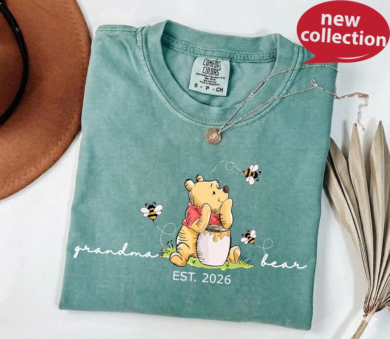 A green t-shirt with a Winnie the Pooh-themed design, featuring the character sitting on a grassy field with a honey pot and a bee nearby. The t-shirt also has the text "grandma bear" and "EST. 2026" printed on it.