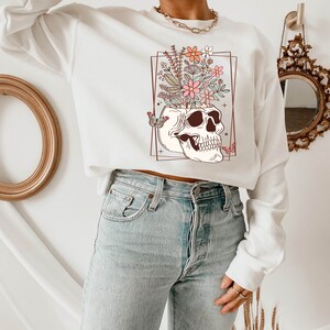 May include: White sweatshirt with a graphic of a skull with flowers growing out of the top. The skull is outlined in brown and the flowers are pink, orange, and white. There are two butterflies on either side of the skull.