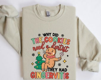 Op de afbeelding: Lichtbeige sweatshirt met de tekst "WHY DID THE COOKIES need a dentist THEY HAD GINGERVITIS" in kleurrijke letters. Een cartoon peperkoekmannetje met een rode sjaal en een lolly staat naast een groene kerstboom.