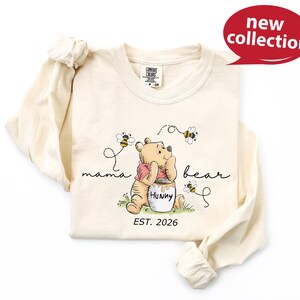 A white long-sleeved t-shirt with a Winnie the Pooh graphic and text, along with a red speech bubble indicating a new collection.