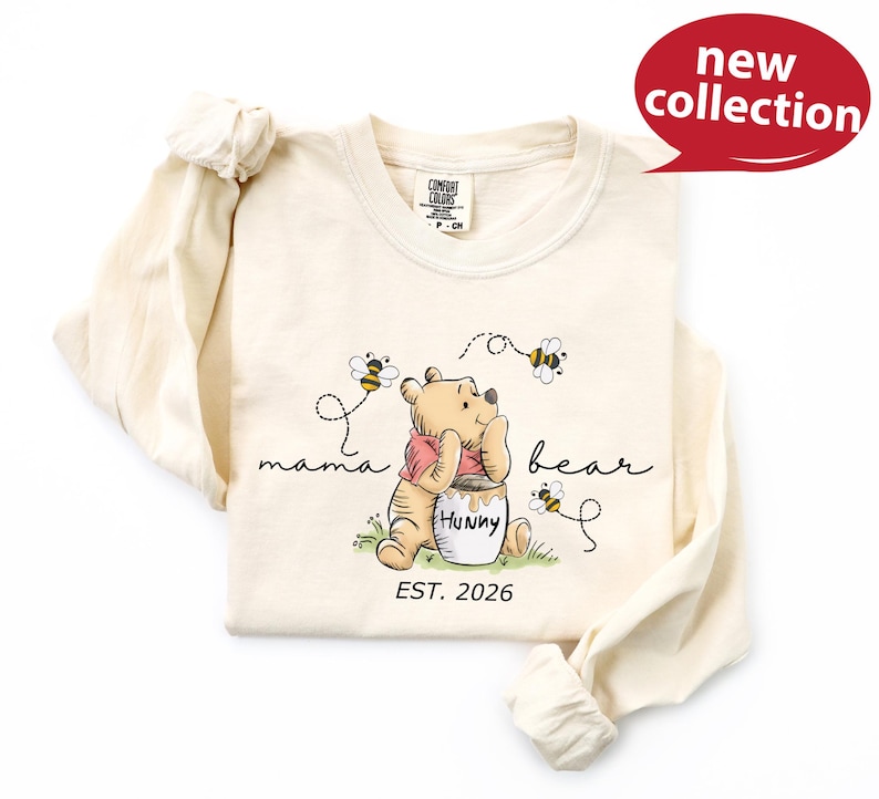 A white long-sleeved t-shirt with a Winnie the Pooh graphic and text, along with a red speech bubble indicating a new collection.