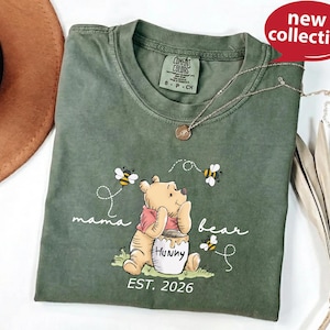 A green t-shirt with a Winnie the Pooh character and the text "mama bear" printed on it, along with the text "Est. 2026". The t-shirt is displayed on a white surface, and there is a brown hat and some leaves in the background.