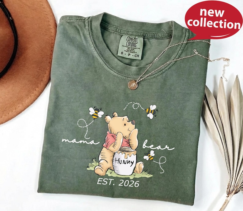 A green t-shirt with a Winnie the Pooh character and the text "mama bear" printed on it, along with the text "Est. 2026". The t-shirt is displayed on a white surface, and there is a brown hat and some leaves in the background.
